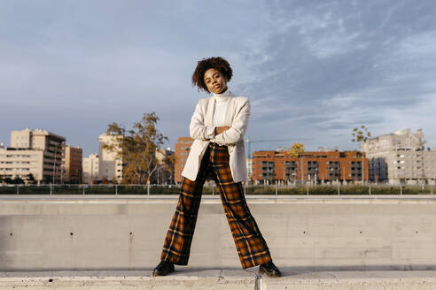 Carefree afro woman with arms crossed posing on retaining wall against buildings in city - TCEF01460