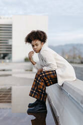 Young woman with afro hair sitting on retaining wall over puddle against building - TCEF01463