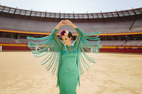 Smiling young woman wearing blue dress and flowers with arms raised dancing in bullring - MIMFF00490