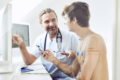 Male shirtless patient with electrodes on chest listening to doctor explaining over computer in hospital stock photo
