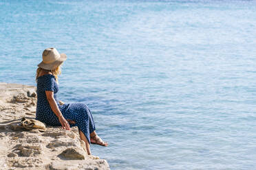 Frau mit Hut schaut auf das Meer, während sie auf einem Felsen sitzt, Menorca, Spanien - DGOF01795