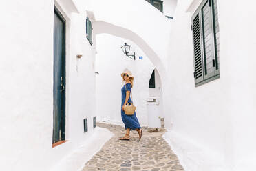 Frau mit Hut geht durch eine Gasse inmitten von Häusern im Dorf Binibeca, Menorca, Spanien - DGOF01802