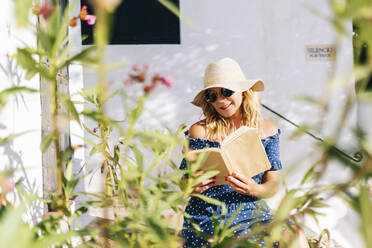 Schöne Frau mit Sonnenbrille liest ein Buch, während sie an der Wand sitzt, Dorf Binibeca, Menorca, Spanien - DGOF01804