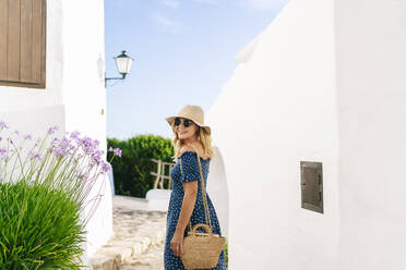 Weiblicher Tourist mit Sonnenbrille und Hut, der über die Schulter schaut, während er im Dorf Binibeca steht, Menorca, Spanien - DGOF01811