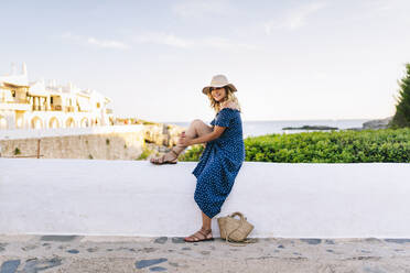 Weibliche Touristin mit Hut sitzt auf einer Stützmauer gegen den Himmel im Dorf Binibeca, Menorca, Spanien - DGOF01825