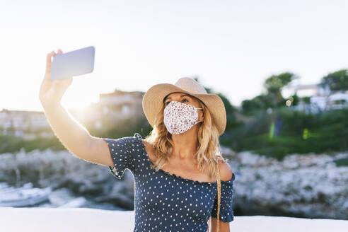 Ältere Frau mit Gesichtsmaske macht ein Selfie mit ihrem Smartphone im Dorf Binibeca, Menorca, Spanien - DGOF01826