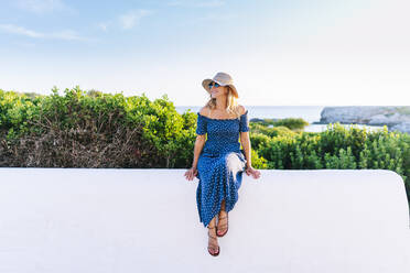 Weibliche Touristin mit Hut sitzt auf einer Stützmauer am Meer im Dorf Binibeca, Menorca, Spanien - DGOF01831