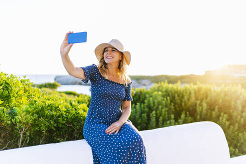 Lächelnde Touristin, die ein Selfie mit ihrem Smartphone vor dem klaren Himmel macht, Dorf Binibeca, Menorca, Spanien - DGOF01833