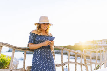 Weibliche Touristin, die ihr Smartphone benutzt, während sie am Geländer gegen den klaren Himmel steht, Dorf Binibeca, Menorca, Spanien - DGOF01834