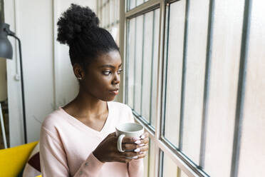 Woman with coffee cup looking through window while standing at home - GIOF10775