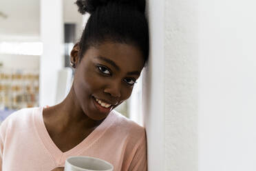 Lächelnde Frau mit Kaffeetasse starrt, während lehnt in der Wand zu Hause - GIOF10780