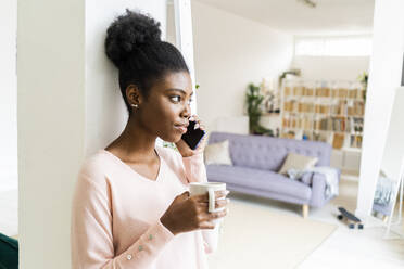 Young woman with coffee cup talking on mobile phone while standing at home - GIOF10781