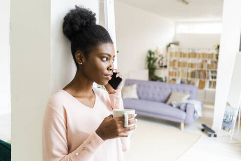 Junge Frau mit Kaffeetasse, die im Stehen zu Hause mit ihrem Handy telefoniert - GIOF10781