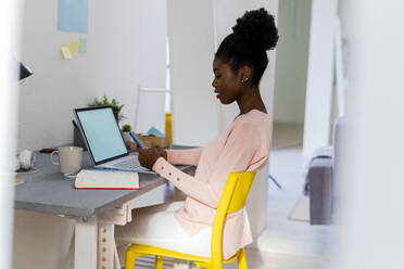 Young businesswoman with laptop using mobile phone while sitting at home office - GIOF10799