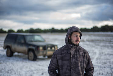 Young man in hooded jacket standing against car at agricultural field - OCMF02004