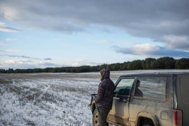 Young man standing by car at agricultural field during winter - OCMF02005