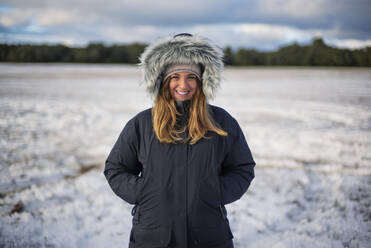 Lächelnde schöne Frau mit Händen in den Taschen, die auf einem verschneiten Feld vor dem Himmel steht - OCMF02008