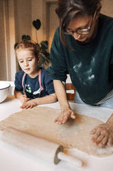 Curious girl watching mother flattening cinnamon rolls dough on table at home - DWF00558