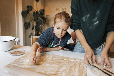 Girl sprinkling cinnamon powder on dough while helping mother in kitchen - DWF00559