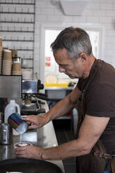 Männlicher Barista in brauner Schürze, der an einer Espressomaschine steht und Milch einschenkt. - MINF15801