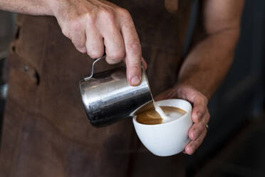 Nahaufnahme eines Barista mit brauner Schürze, der Cappuccino einschenkt. - MINF15808