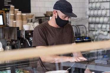 Männlicher Barista mit schwarzer Baseballkappe und Gesichtsmaske arbeitet hinter dem Tresen. - MINF15815