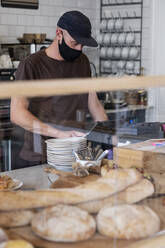 Männlicher Barista mit schwarzer Baseballkappe und Gesichtsmaske arbeitet hinter dem Tresen. - MINF15816