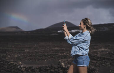 Junge Frau, die ein Foto mit ihrem Mobiltelefon macht, während sie am Vulkan El Cuervo steht, Lanzarote, Spanien - SNF00956