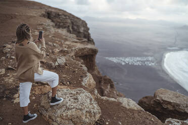 Weibliche Touristin, die ein Foto mit ihrem Handy macht, während sie auf einem Berg am Strand von Famara steht, Lanzarote, Spanien - SNF00983