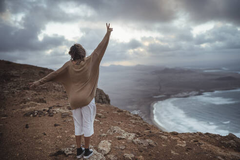 Verspielte Frau mit Friedenszeichen auf einem Berg am Strand von Famara, Lanzarote, Spanien - SNF00988