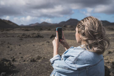 Junge Frau, die ein Foto mit ihrem Mobiltelefon macht, während sie am Vulkan El Cuervo steht, Lanzarote, Spanien - SNF00996