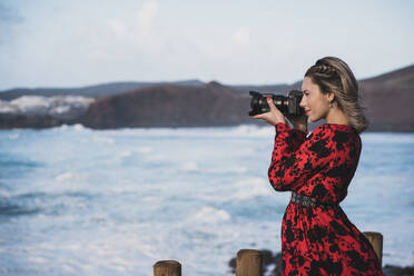 Young woman photographing with camera while standing against sea at El Golfo, Lanzarote, Spain - SNF01015