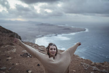 Lächelnde Frau, die mit ausgestreckten Armen auf einem Berg vor dem Strand von Famara steht, Lanzarote, Spanien - SNF01016
