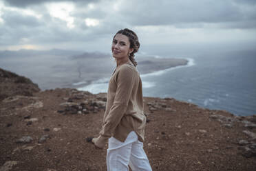 Young woman smiling while walking on mountain against Famara Beach, Lanzarote, Spain - SNF01017