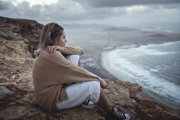 Junge Frau schaut auf den Strand von Famara, während sie auf einem Berg in Lanzarote, Spanien, sitzt - SNF01020