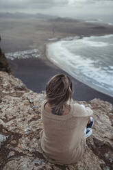Weibliche Forscherin betrachtet die Aussicht, während sie auf einem Berg gegenüber dem Strand von Famara sitzt, Lanzarote, Spanien - SNF01021
