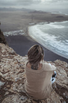 Female explorer looking at view while sitting on mountain against Famara Beach, Lanzarote, Spain - SNF01021