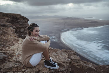 Lächelnde Frau, die ihre Knie umarmt, während sie auf einem Berg vor dem Strand von Famara sitzt, Lanzarote, Spanien - SNF01024