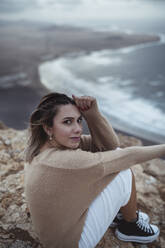 Frau, die auf einem Berg sitzend den Strand von Famara anstarrt, Lanzarote, Spanien - SNF01025