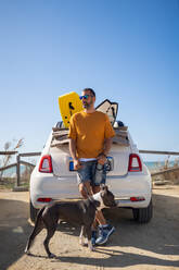 Männlicher Besitzer mit Pitbull-Hund, der an einem sonnigen Tag am Strand in der Nähe des Autos steht und Spaß hat - ADSF20190