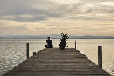 Back view of male biker sitting on wooden quay with parked motorbike and enjoying sunset over sea in evening - ADSF20197