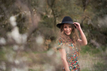 Side view of delighted female with long hair and in dress standing in woods while touching hat and looking at camera - ADSF20201