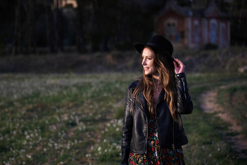 Stilvolle Frau mit Hut und schwarzer Lederjacke steht bei Sonnenuntergang auf einem Feld und schaut weg - ADSF20205