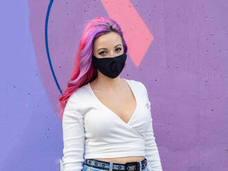 Modern millennial hipster female with dyed long hair wearing black protective mask for coronavirus prevention looking at camera while standing against bright colorful wall - ADSF20230