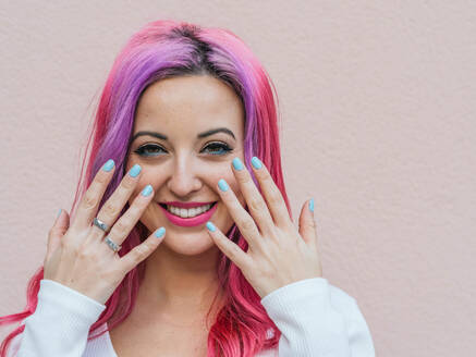 Lächelnde junge Frau mit rosa Haaren und hellem Make-up zeigt Hände mit blau gefärbten Nägeln vor beigem Hintergrund - ADSF20232