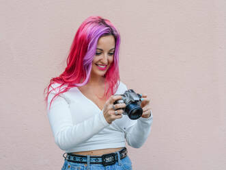Junge Hipster-Frau mit rosa Haaren schaut in die Kamera, während sie ein Foto vor einer beigen Wand schießt - ADSF20233
