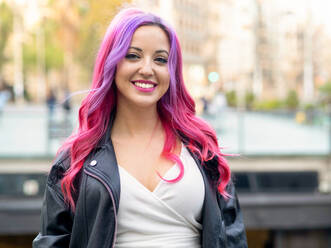 Optimistische, selbstbewusste Millennial-Frau mit rosa gefärbtem Haar und Lederjacke, die in die Kamera schaut und lächelt, während sie vor einem unscharfen städtischen Hintergrund steht - ADSF20235