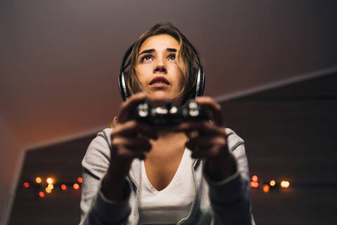 From below concentrated female in headphones and with console sitting in armchair and playing video game while entertaining in evening at home stock photo