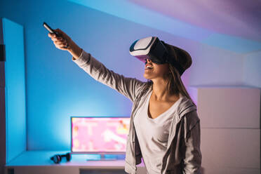 Aufgeregte weibliche Spielerin spielt ein Videospiel in einem Virtual-Reality-Headset, während sie in einem Raum mit Neonlicht sitzt - ADSF20253