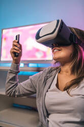 Aufgeregte weibliche Spielerin spielt ein Videospiel in einem Virtual-Reality-Headset, während sie in einem Raum mit Neonlicht sitzt - ADSF20254